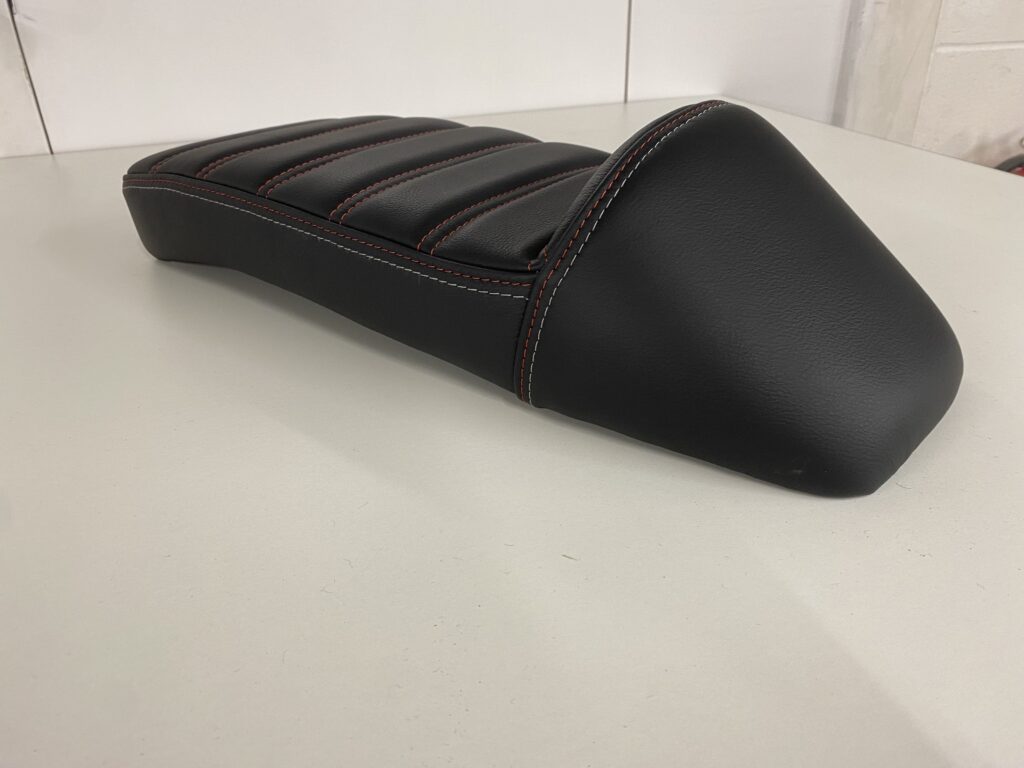 A Lambretta style, long bike seat, black in colour with fresh red stitching.